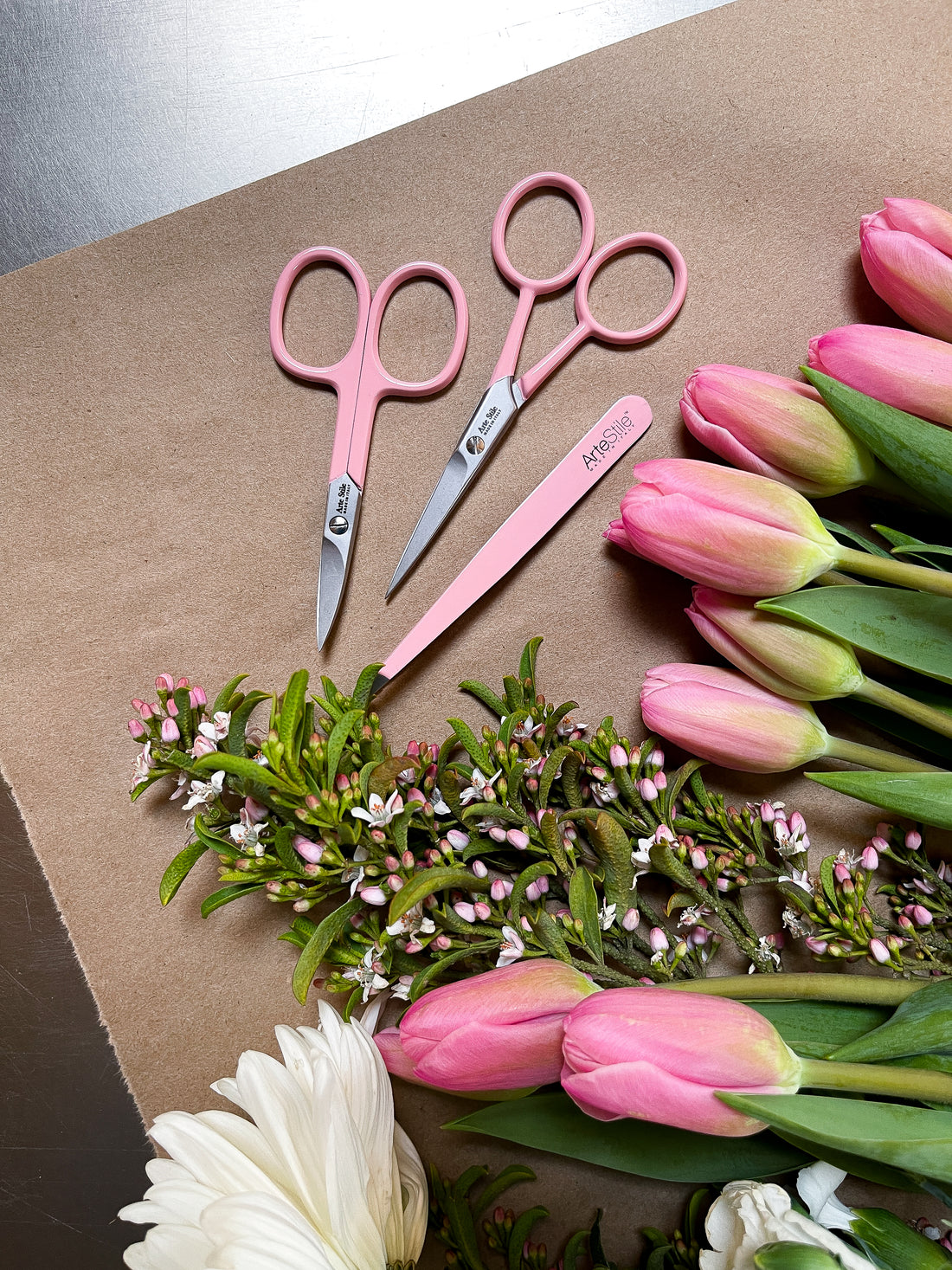 ArteStile Brow Scissors, ArteStile Nail Scissors, and ArteStile tweezers in rosé (light pink), on a brown wood table with pink tulips. 