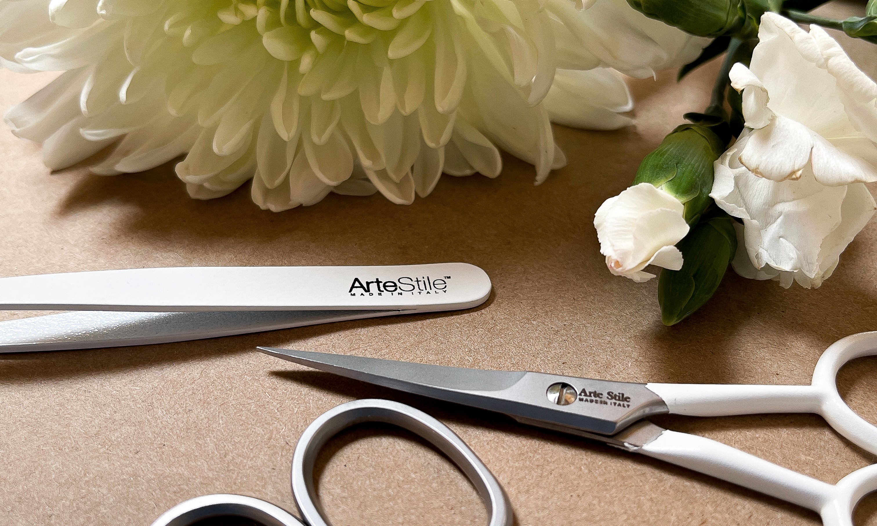 A pair of tweezers and two fairs of scissors on a beige background. There are several white flowers in frame.