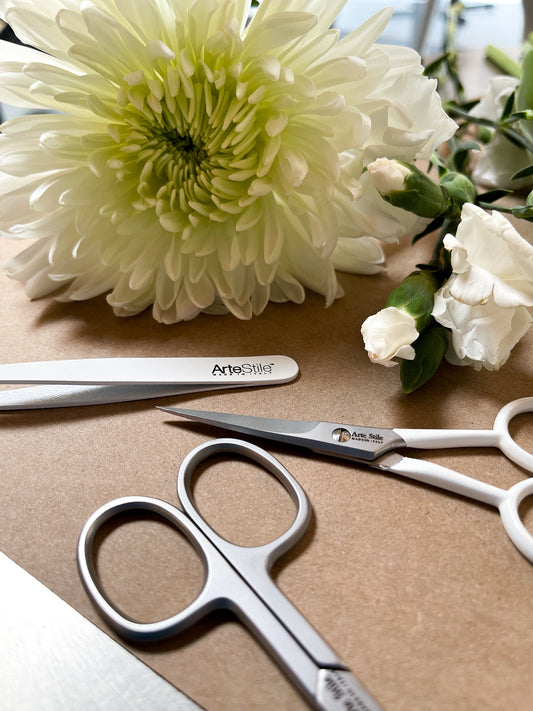 A pair of tweezers and two fairs of scissors on a beige background. There are several white flowers in frame.